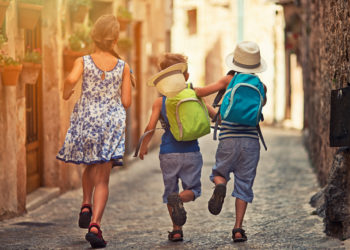 Three kids running and having fun in the beautiful mediterranean town. Little boys are wearing backpacks and hats. One hat is falling off the boy's head. Theirs sister is wearing sundress.