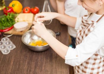 Small teenage girl in apron mixing eggs in white bowl in the kitchen