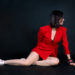 Young beautiful wearing short bright red dress and white sneakers, sits on the floor on black background.. Dark hair, slim shape, long legs, face sideways, copy space.